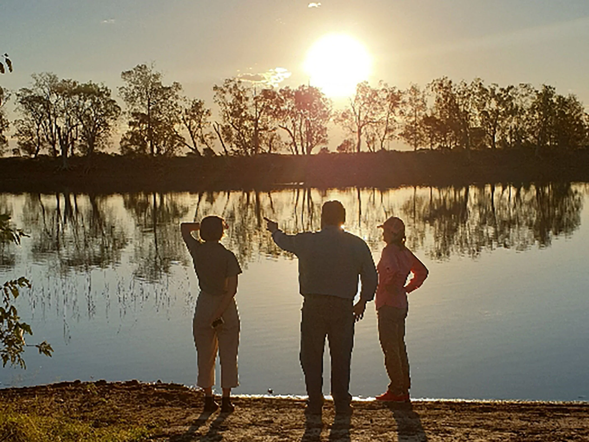 Three people standing on a riverbank at sunset with trees reflecting in the water, one person pointing towards the horizon.
