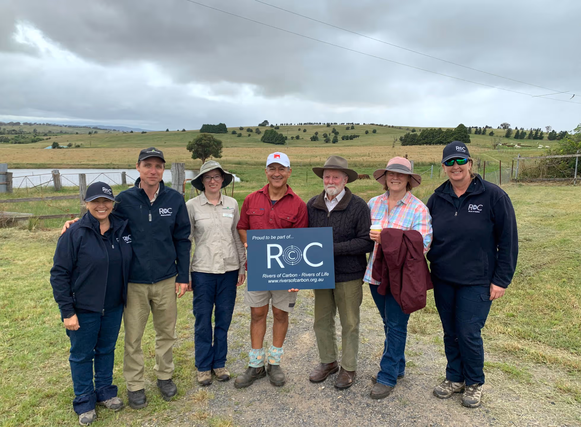 Group of seven people standing outdoors in a grassy rural area, some wearing hats and holding a sign that reads 'Proud to be part of... ROC Rivers of Carbon - Rivers of Life www.riversofcarbon.org.au'.