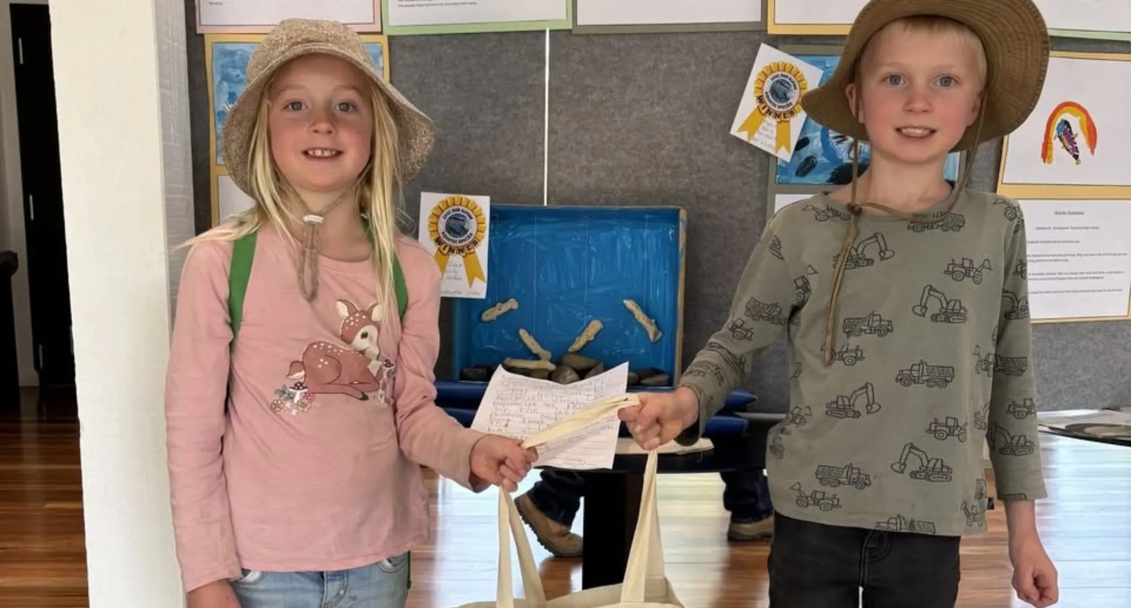 Two children wearing sun hats and holding a paper and a beige tote bag, standing indoors near a display board with ribbons and artwork.
