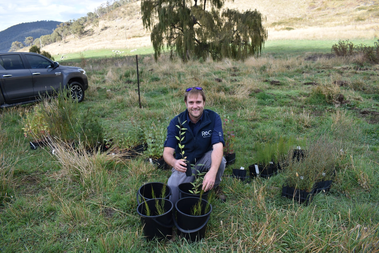 Man kneeling in grass holding a small plant, surrounded by pots and seedling trays in a rural field with a parked truck nearby.
