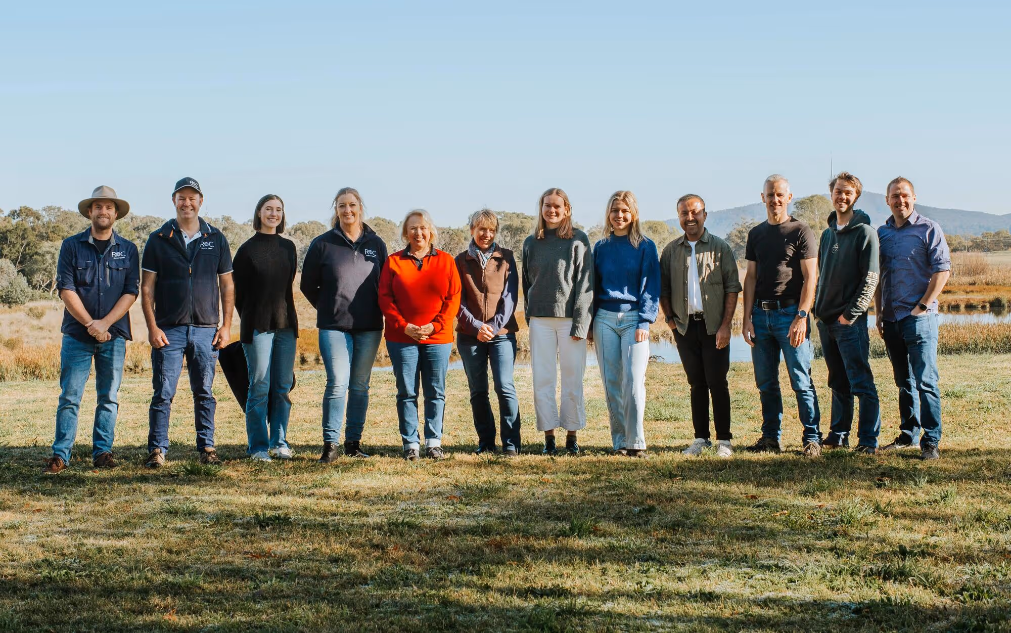 Group of thirteen people standing outdoors on grass with trees and hills in the background.