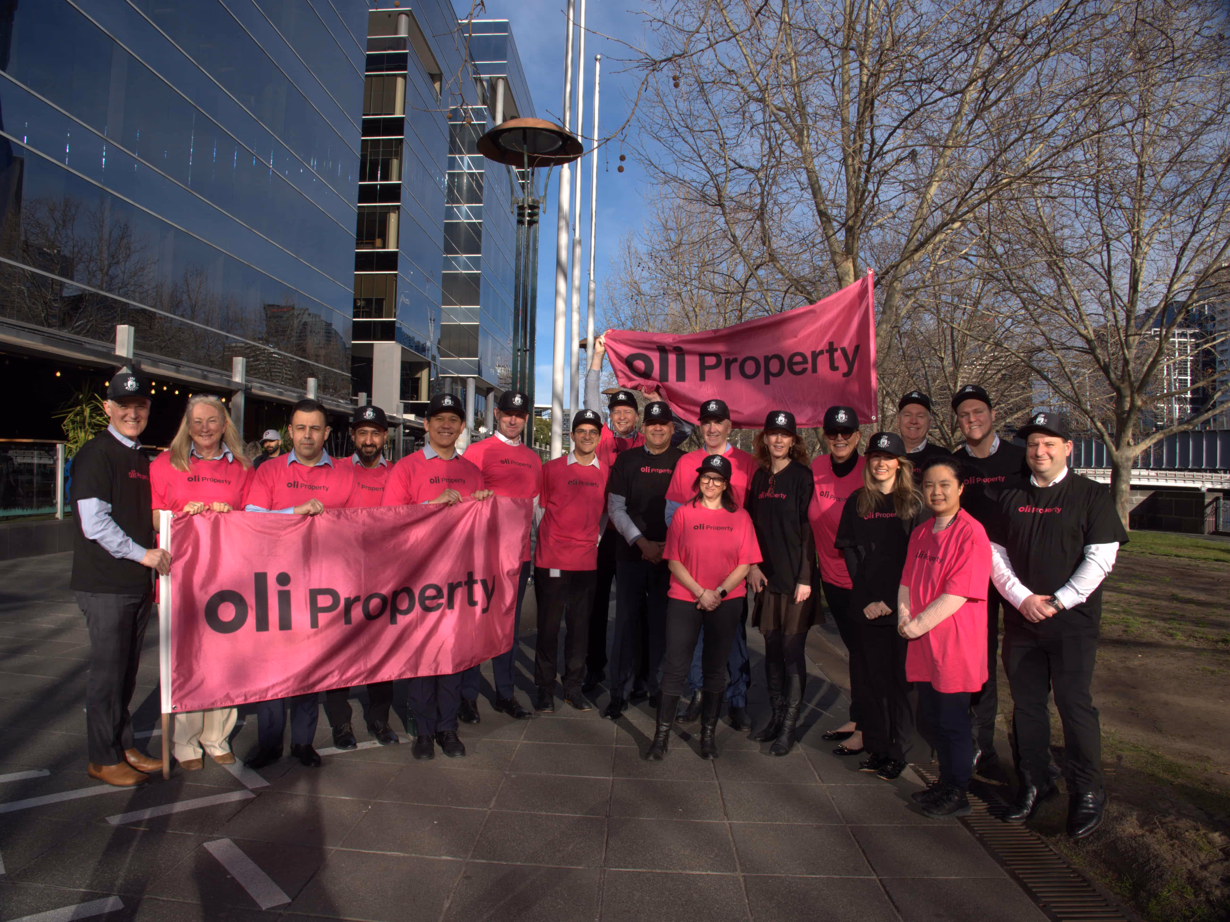 Group of people wearing black and pink oli Property shirts and caps, holding two pink oli Property banners outdoors near modern buildings and leafless trees.