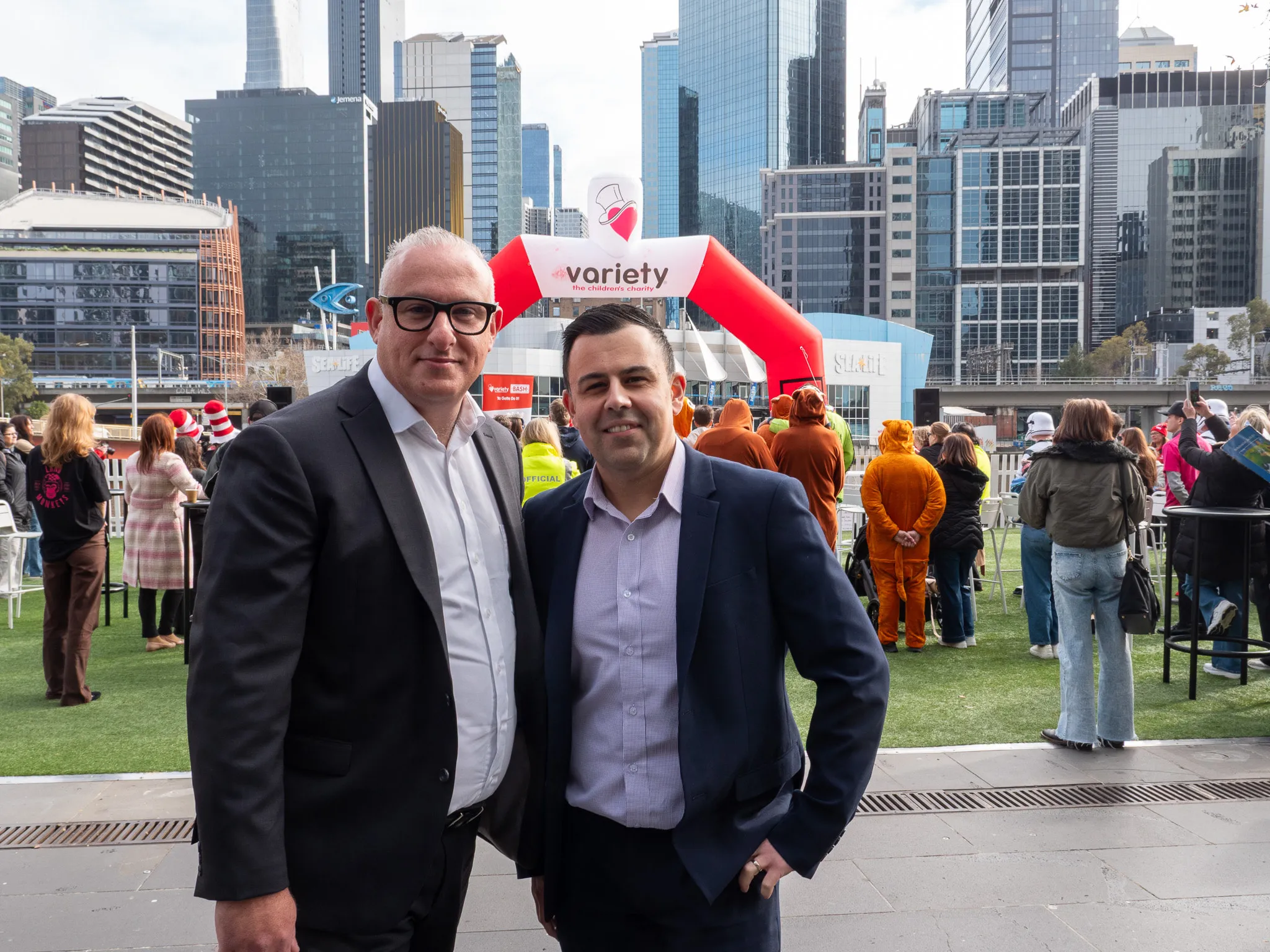 Two men in suits smiling and posing at an outdoor charity event with city skyscrapers in the background and people in animal costumes.