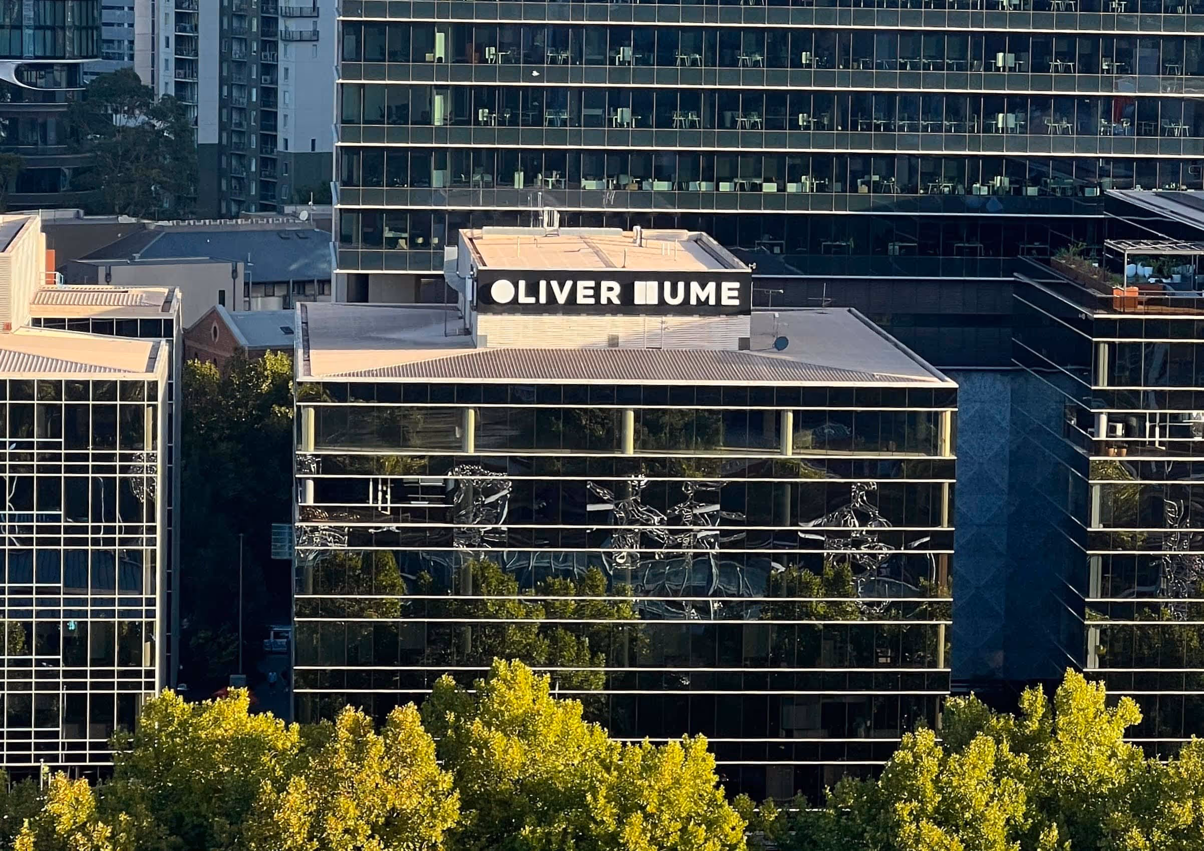 Modern office building with reflective glass windows and a rooftop sign partially reading 'OLIVER MUME', surrounded by trees and other buildings.
