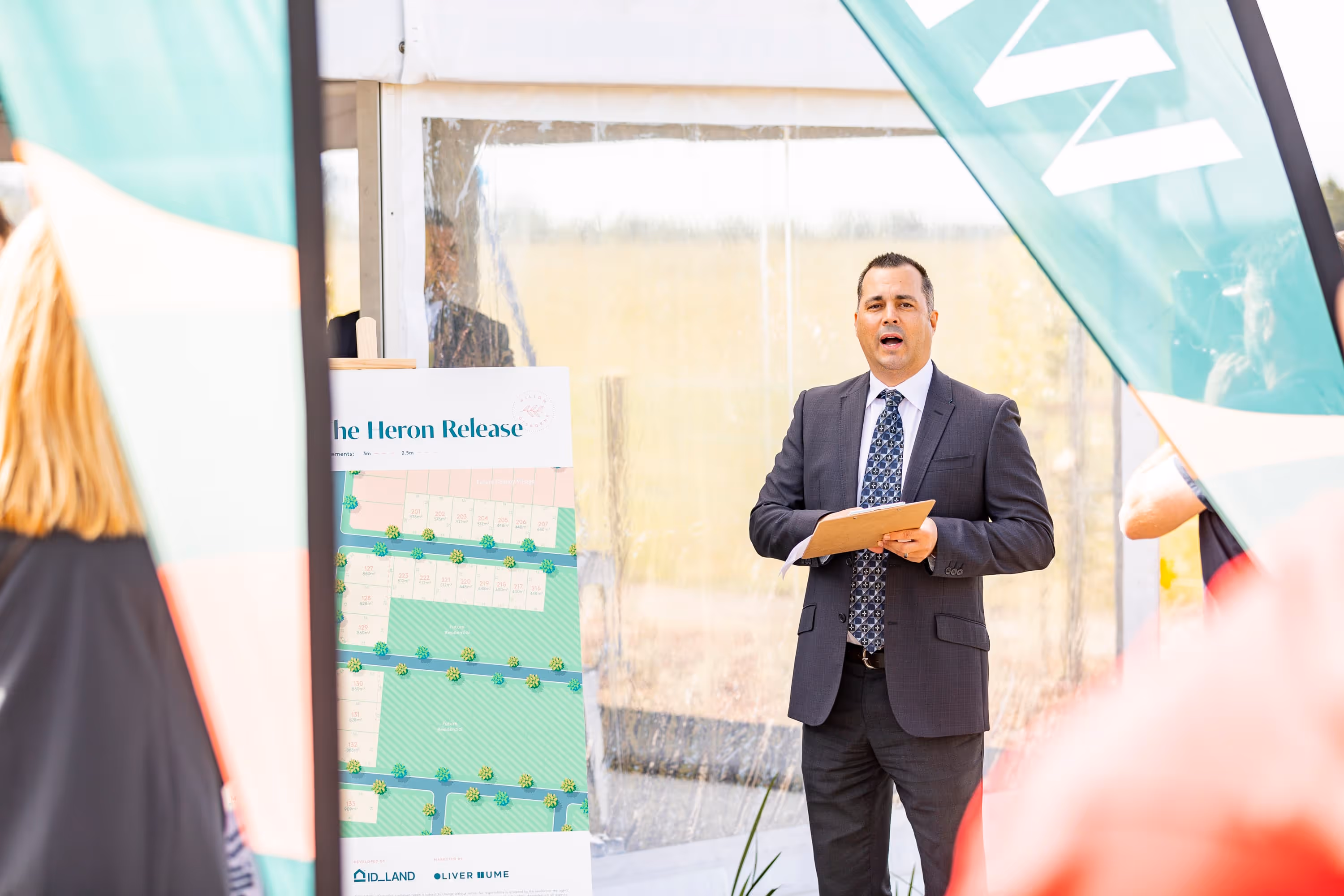 Man in a suit holding a clipboard speaking at an outdoor event next to a display board titled 'The Heron Release'.