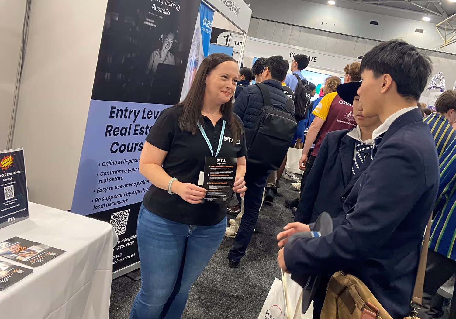 A woman at a real estate training booth talks to two young men in suits at a crowded event.