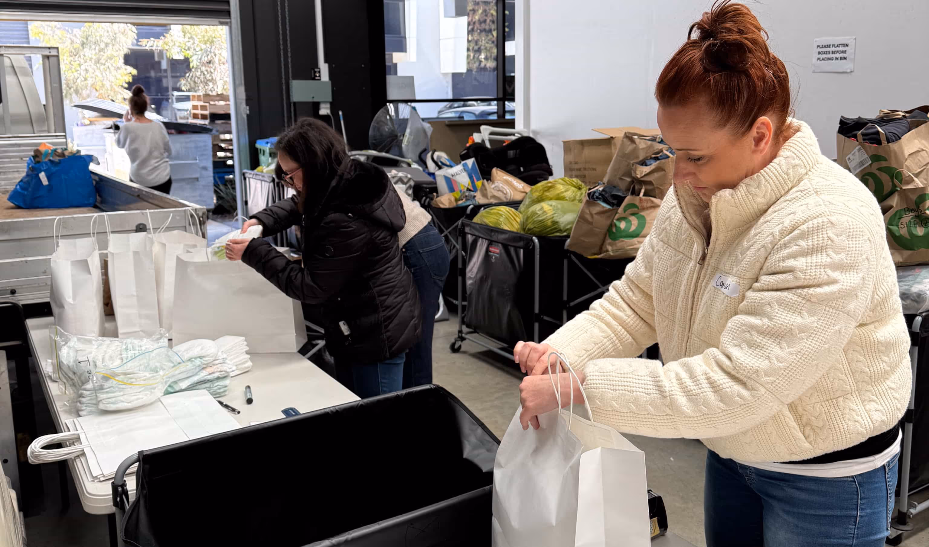 Three women packing white paper bags with groceries and supplies in a warehouse setting.