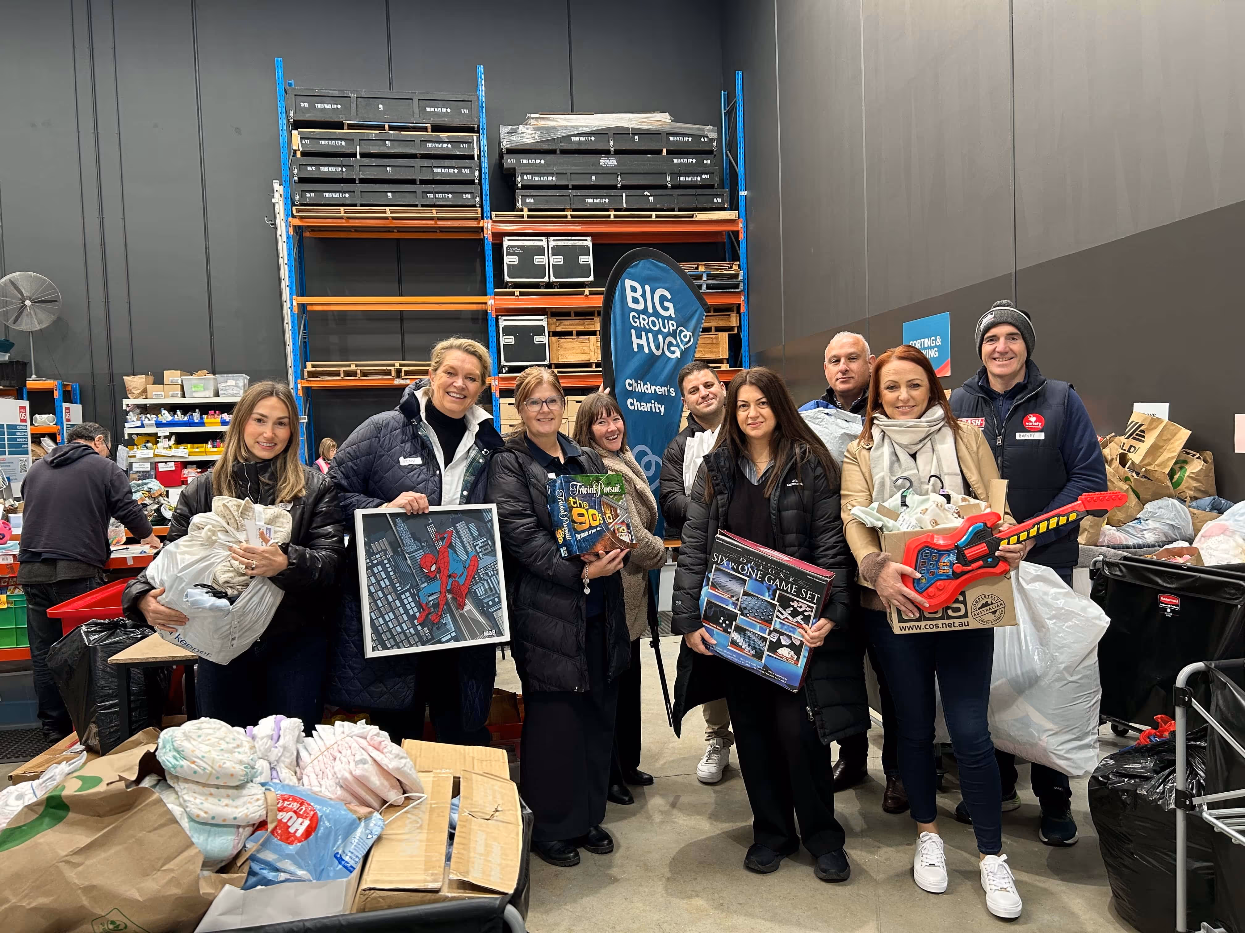 Group of eight people in a warehouse holding toys and games in front of a Big Group Hug Children's Charity banner.