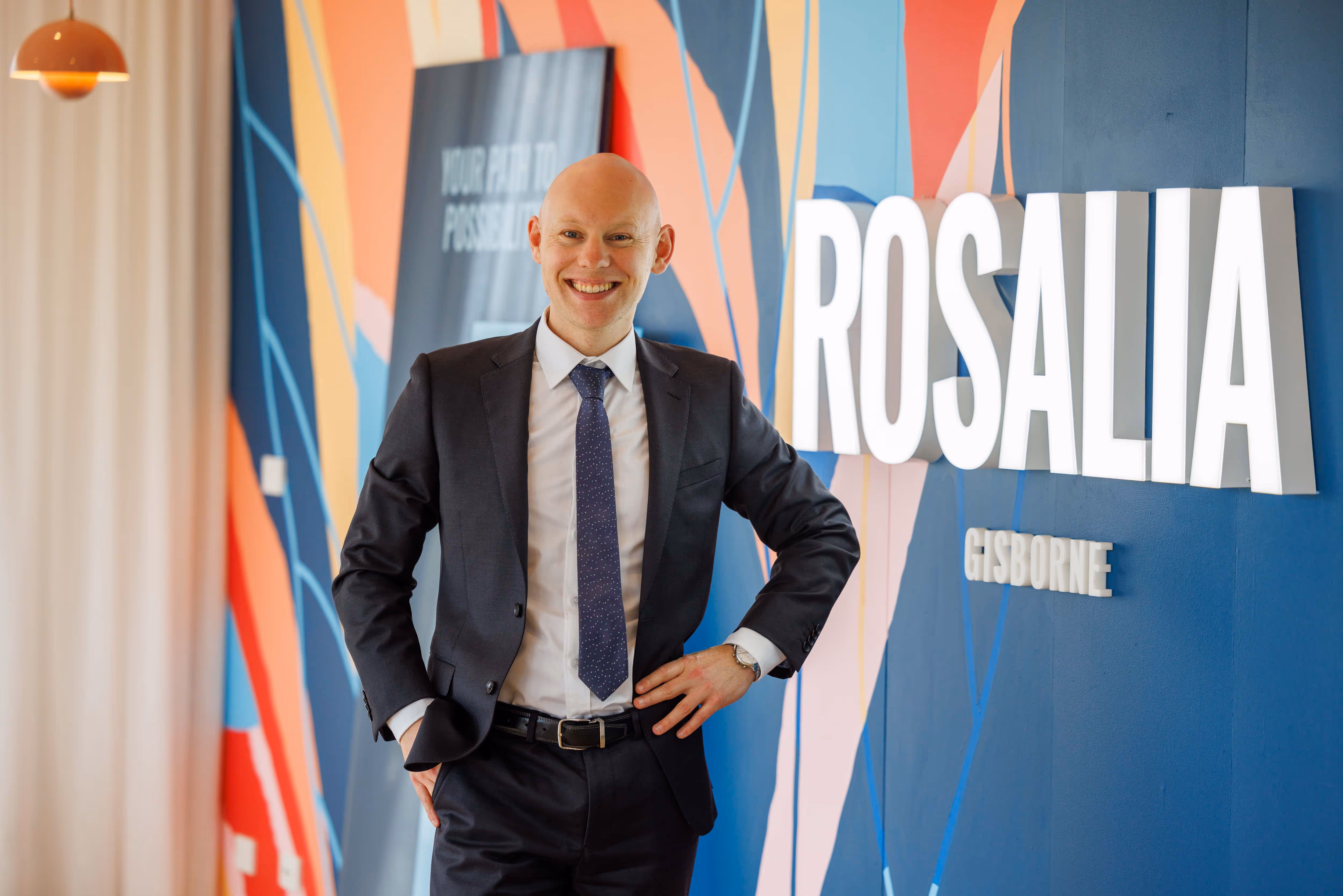 Smiling bald man in a dark suit and tie standing in front of a colorful mural with the text 'ROSALIA GISBORNE'.