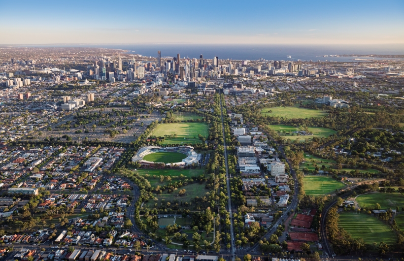 Aerial view of Melbourne, showing the city skyline, Albert Park’s green open spaces, tree-lined boulevards, and a sports stadium in the foreground, with Port Phillip Bay visible in the distance under a clear blue sky.