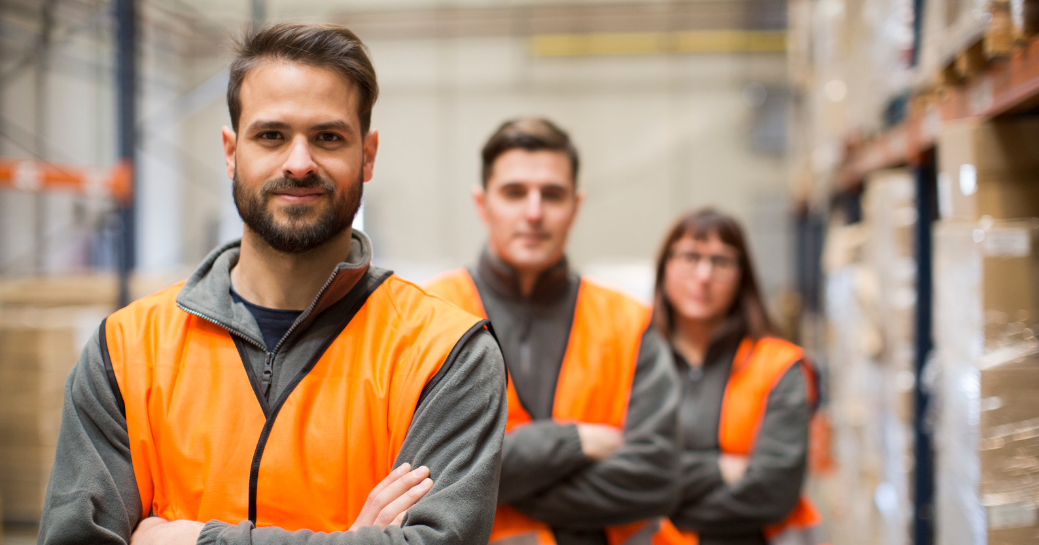 Three men in bright orange vests are positioned in a warehouse, observing their surroundings filled with storage racks and tools.
