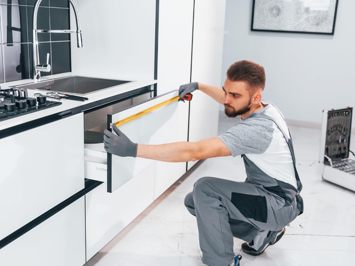 Man in gray work overalls measuring kitchen cabinet door with a tape measure.