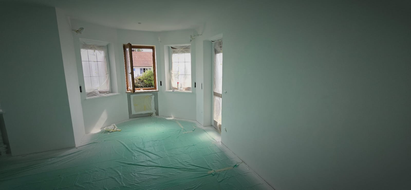 Empty room with white walls and windows covered in protective plastic, floor covered with green plastic sheet for painting preparation.