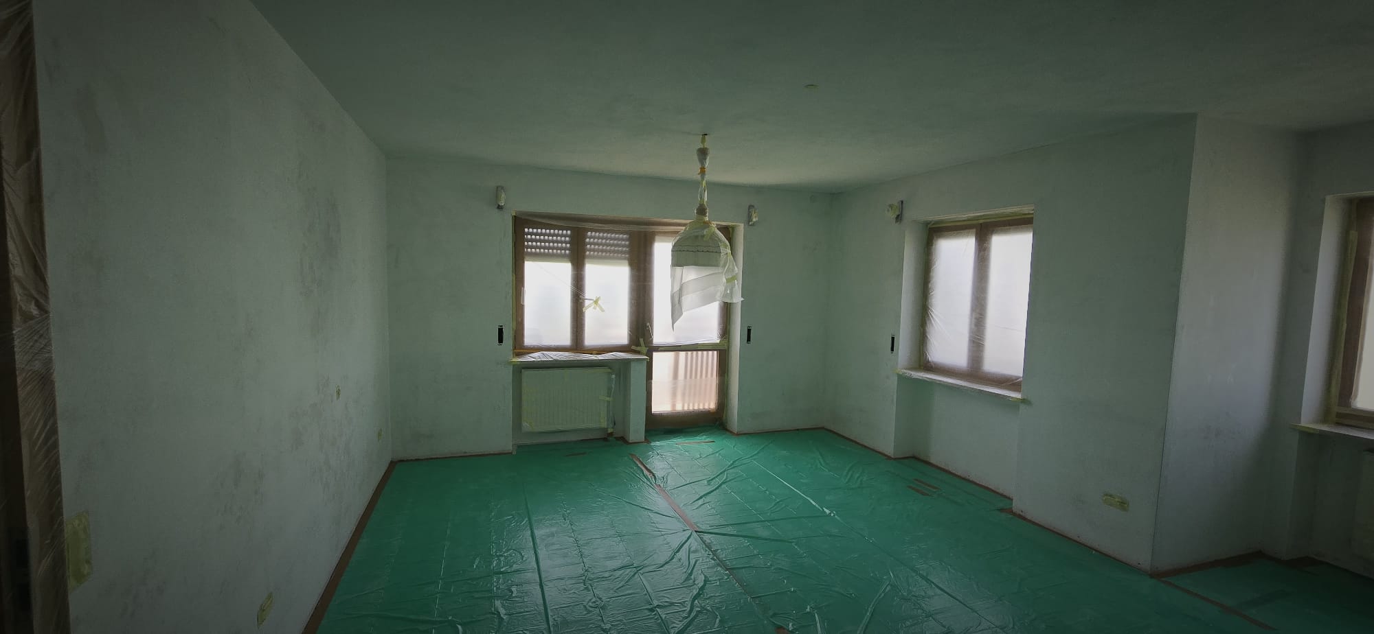Empty room with windows covered in plastic and floor covered with green protective sheet, walls showing signs of wear or mold.