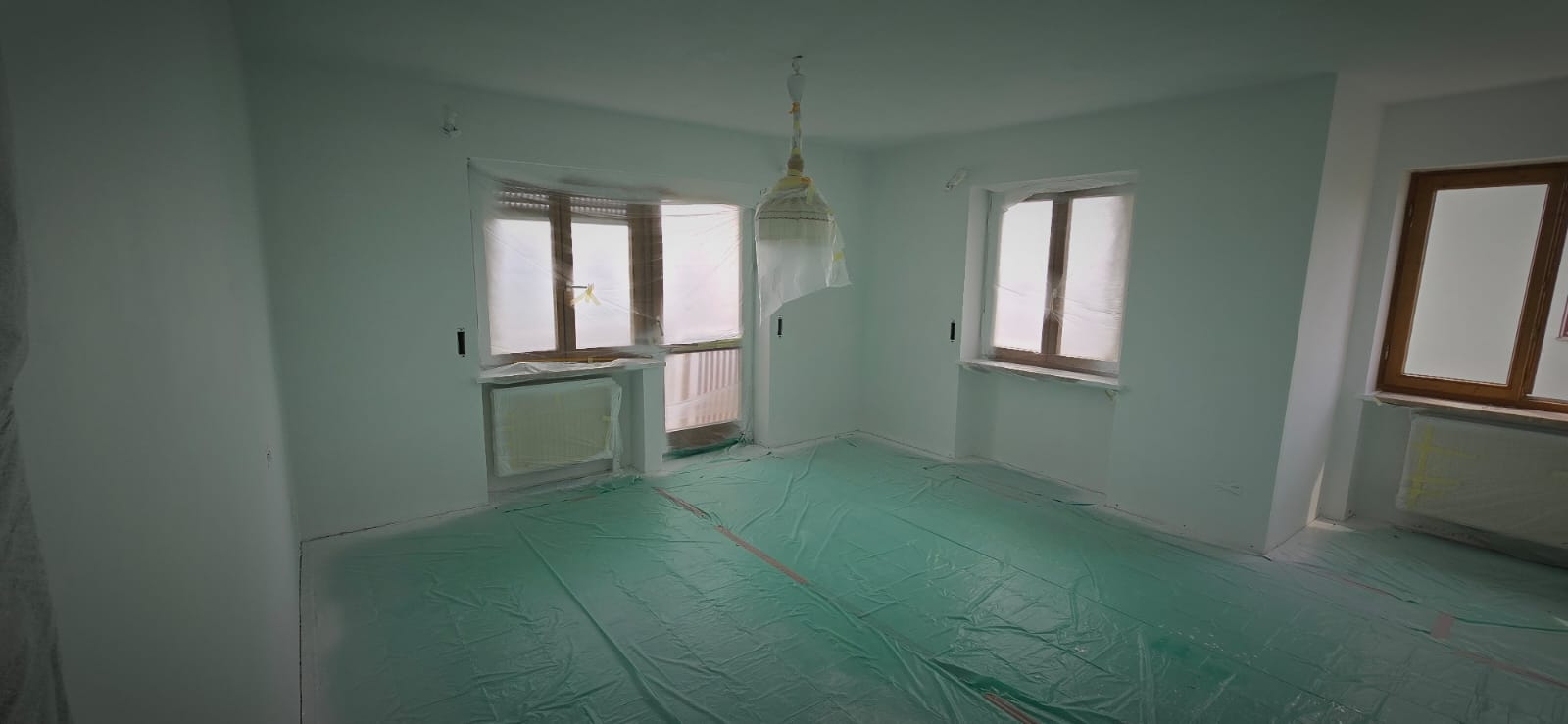 Empty room with windows and door covered in plastic sheets and floor protected with green plastic during painting.