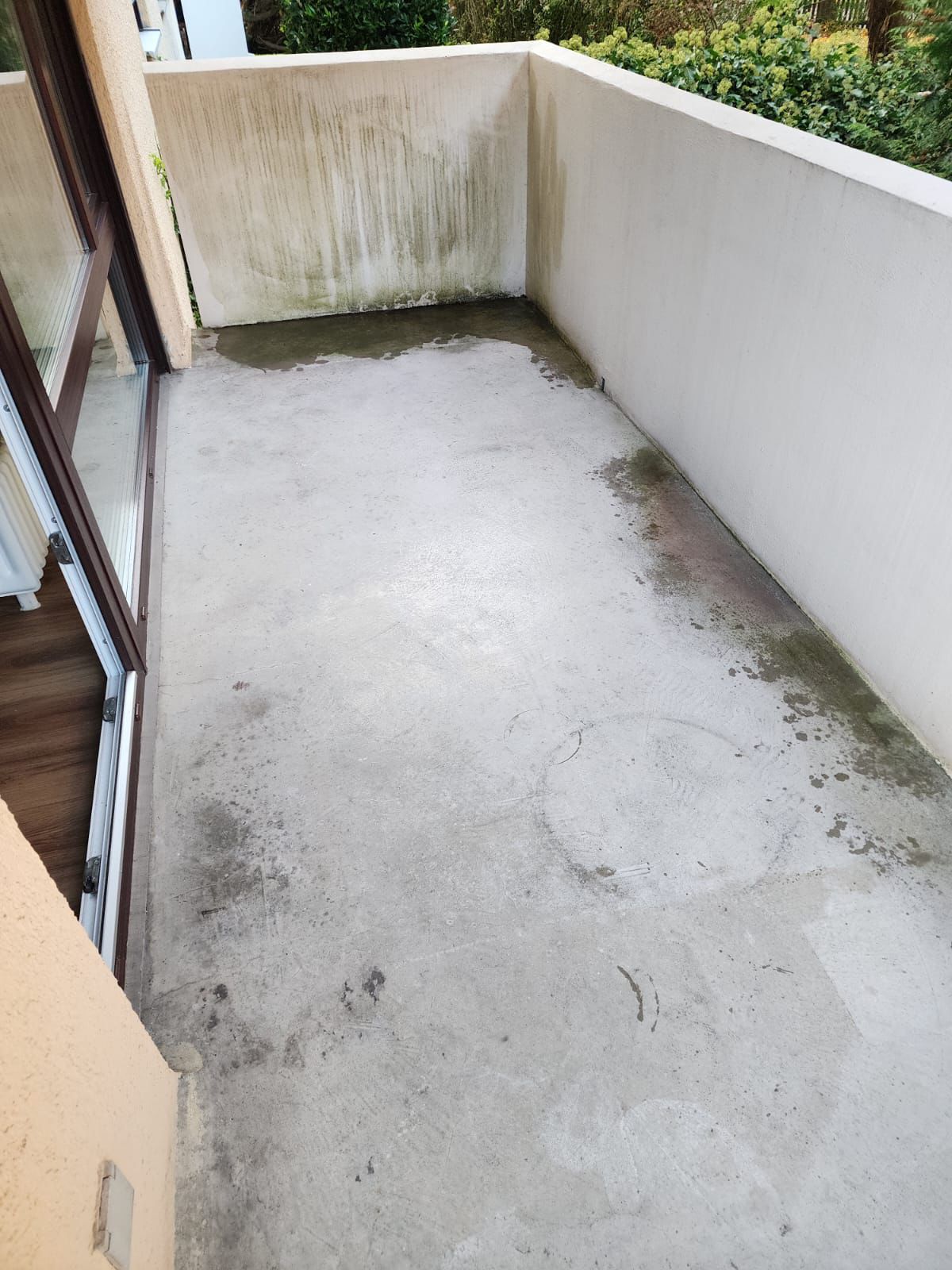 Concrete balcony floor with visible stains, dirt, and damp marks near the walls, next to a glass door leading inside.
