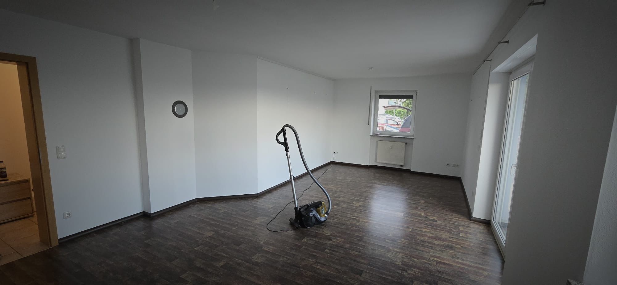Empty room with dark wood flooring, white walls, a window, a glass door, and a vacuum cleaner in the center.
