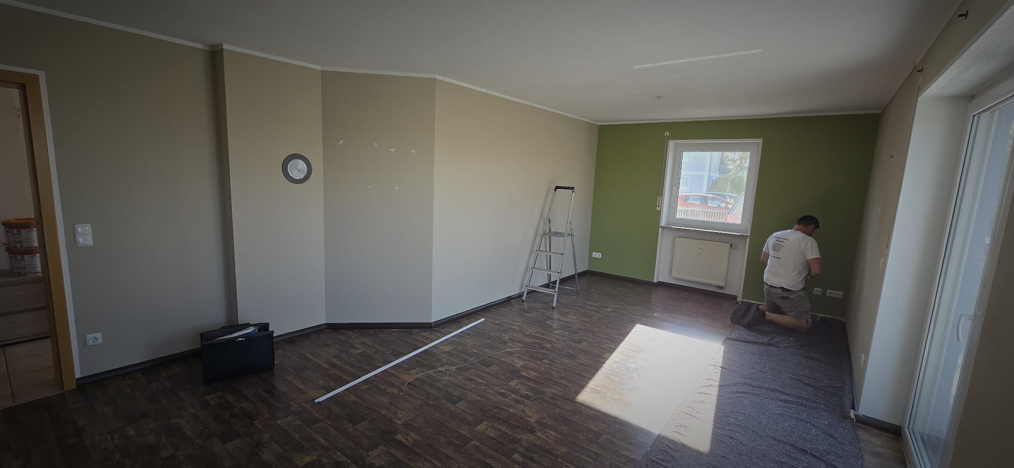 Man kneeling on floor in a room with green and beige walls, wooden floor, a ladder, and sunlight coming through a window.