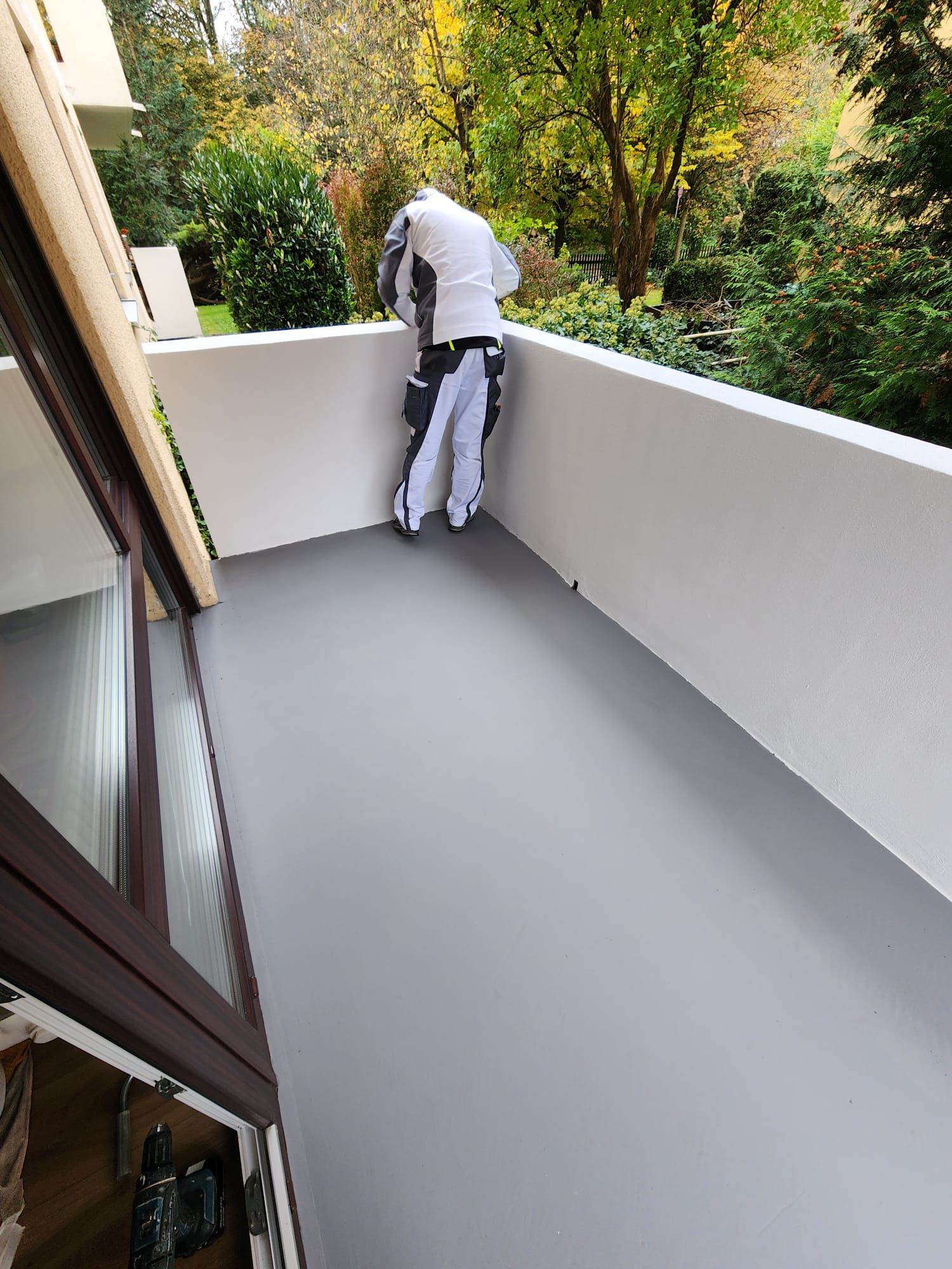 Person in work attire standing on a freshly painted gray balcony with white walls, surrounded by green trees.