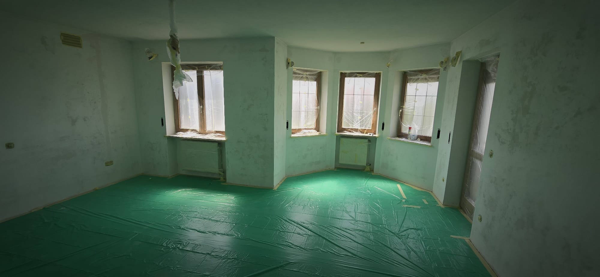 Empty room under renovation with plastic-covered windows and green protective floor covering.