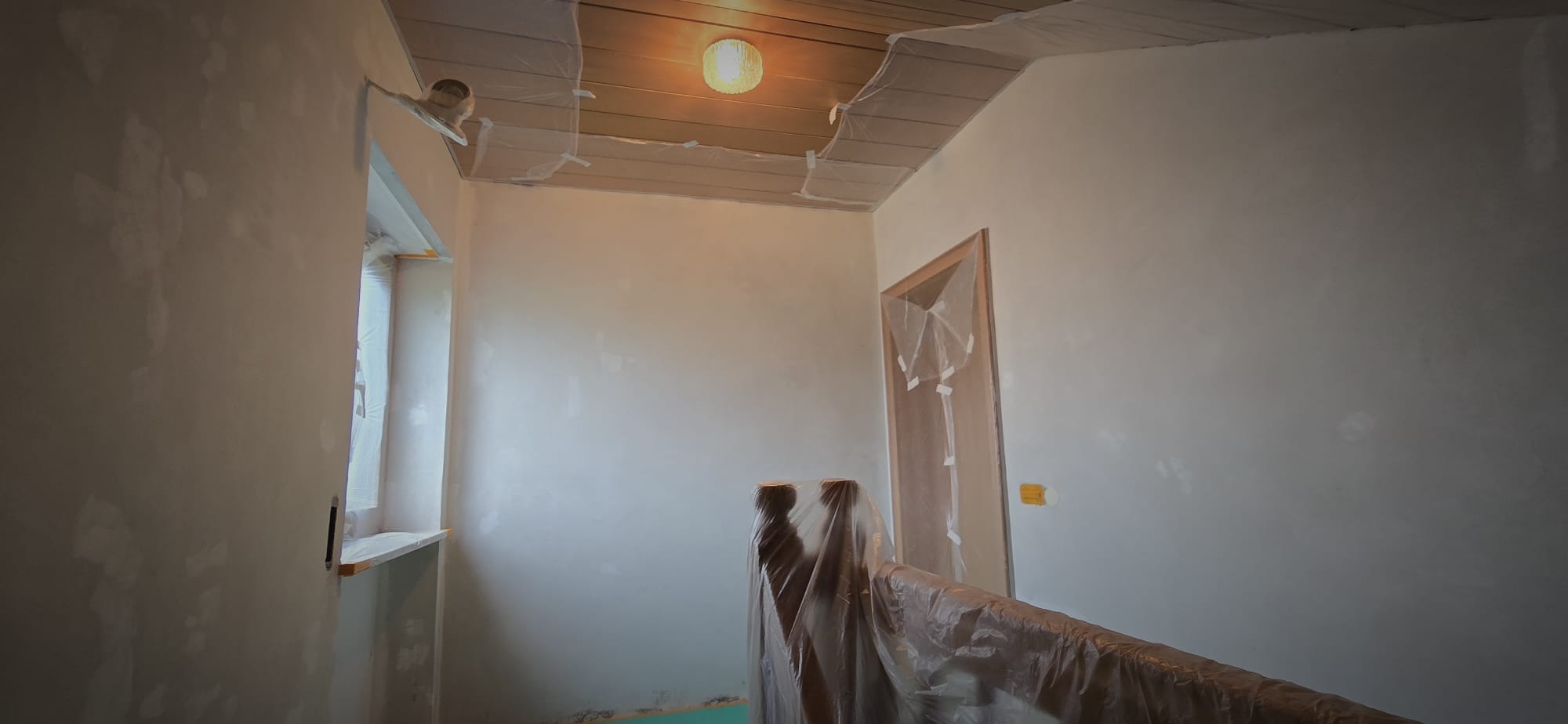 Interior room under renovation with white walls, a covered door and window, and plastic-covered furniture and banister.