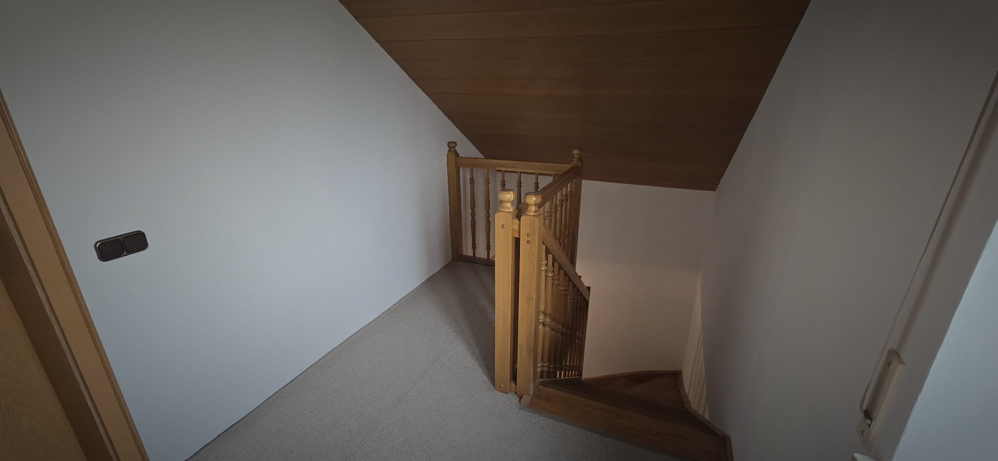 Top of wooden staircase with brown handrails, white walls, gray floor, and angled wooden ceiling above.