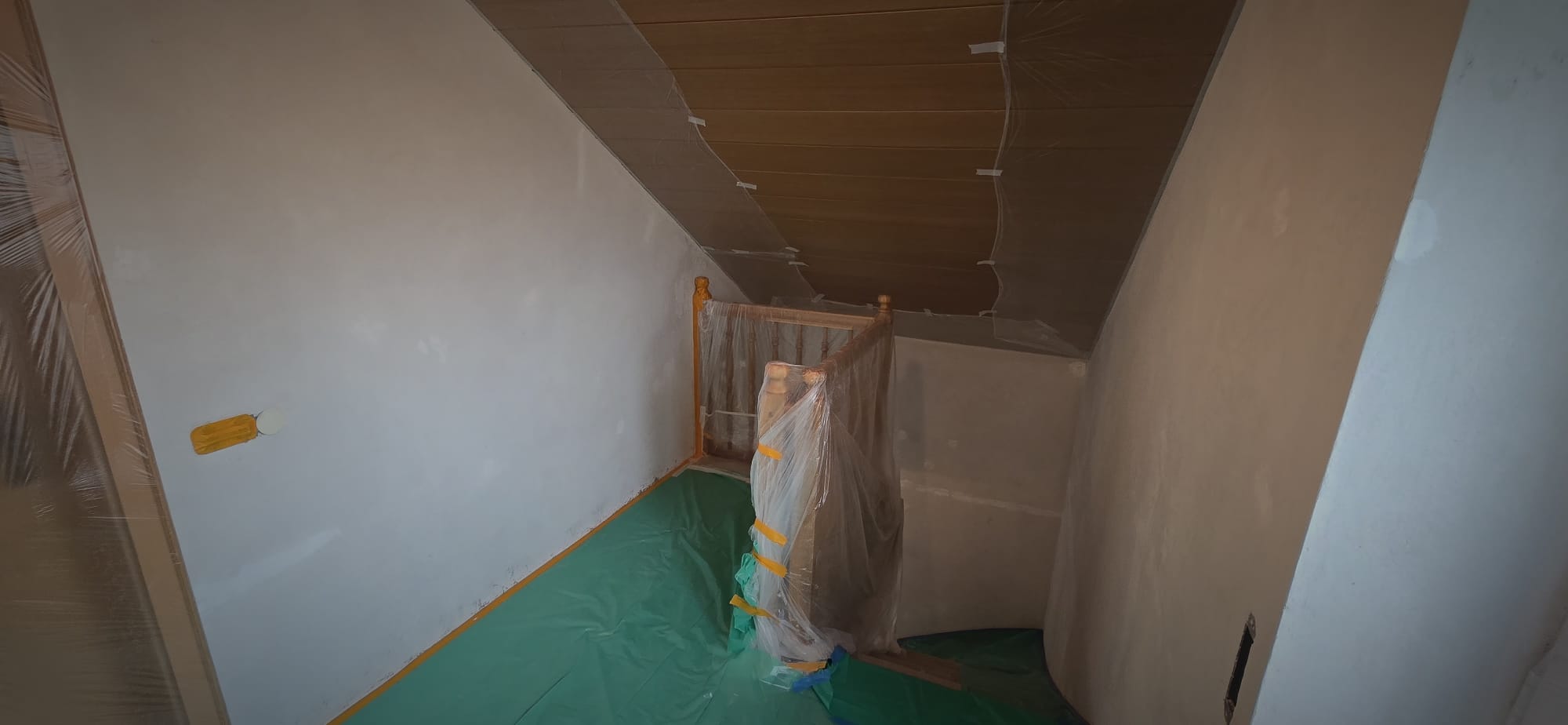 Staircase area with wooden railing covered in protective plastic and green floor covering, walls partially painted.