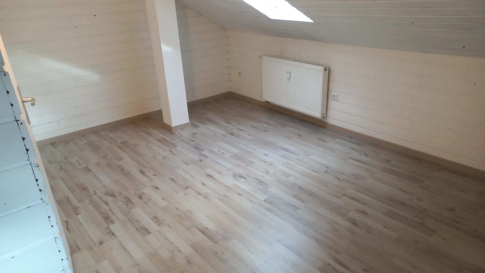 Empty attic room with light wood flooring, white paneled walls, a radiator, and a slanted ceiling with a skylight.