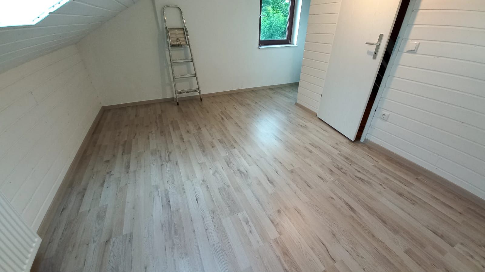 Empty room with light wood laminate flooring, white paneled walls, a window, an open door, and a metal folding ladder leaning against the wall.