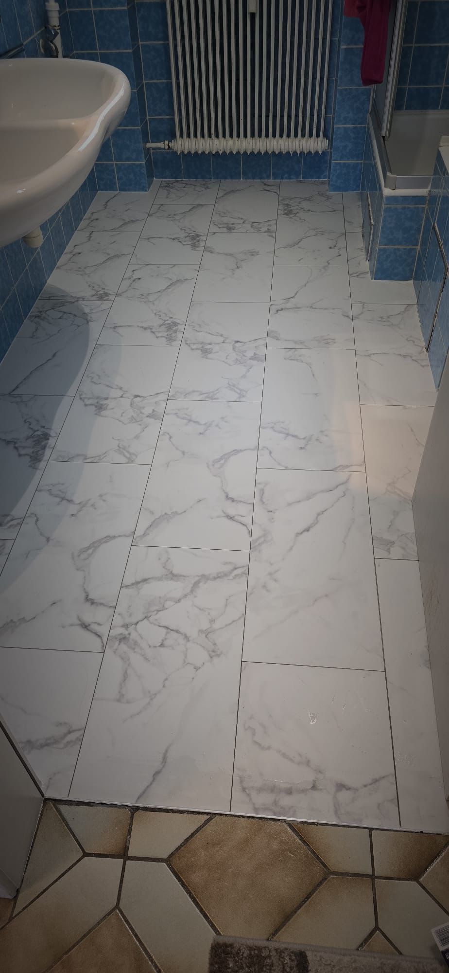 Bathroom floor with newly installed white marble-effect tiles transitioning from beige hexagonal tiles, blue tiled walls, white sink, and radiator.