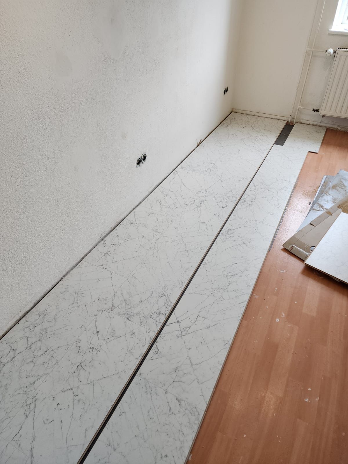 Partially installed white marble-patterned floor panels in a room with light wooden flooring and white walls.