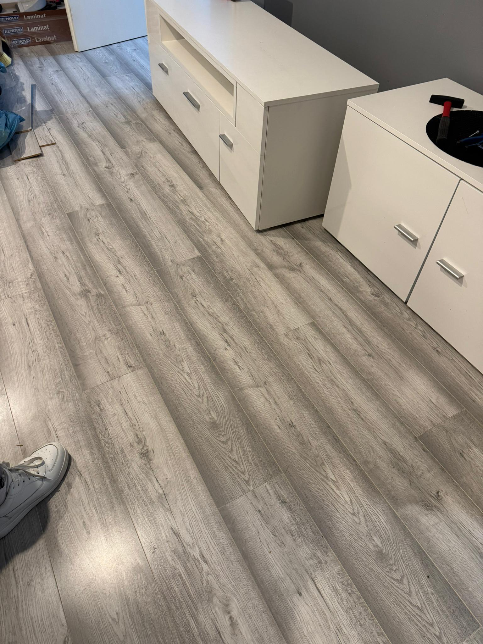 Newly installed light gray wood laminate flooring in a room with white cabinets and a person's white sneaker visible.