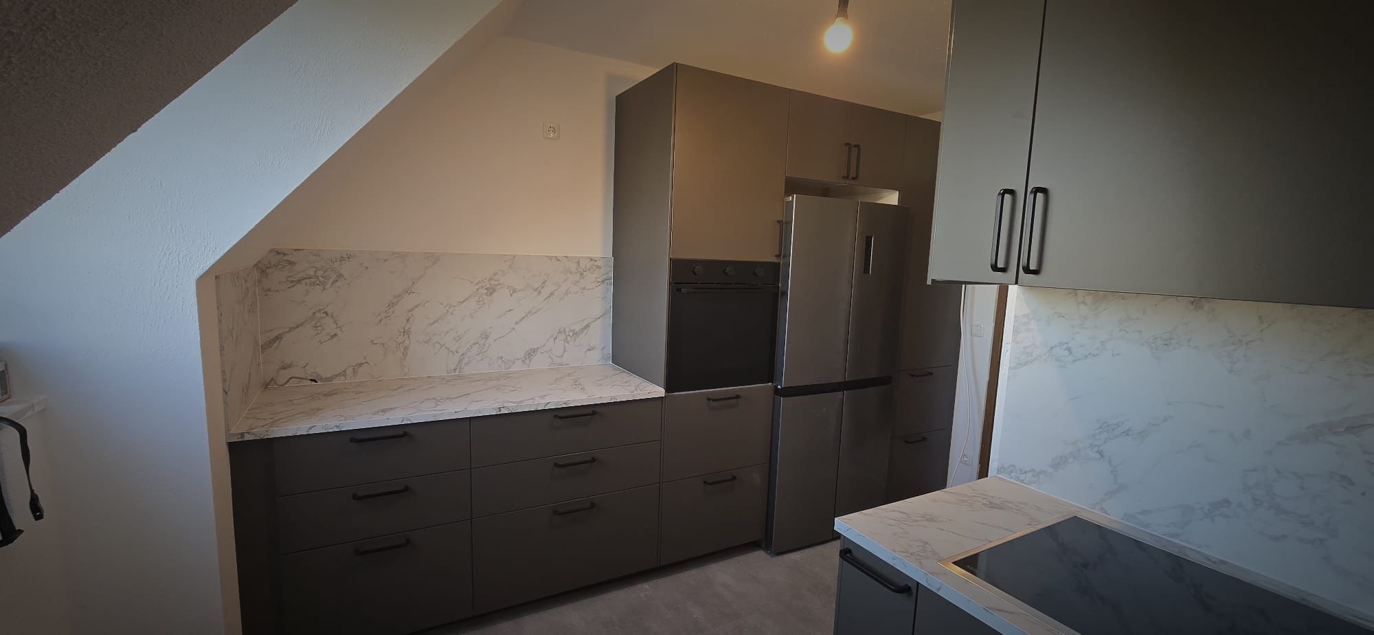 Modern kitchen with gray cabinets, marble countertops and backsplash, built-in oven, and stainless steel refrigerator under slanted ceiling.