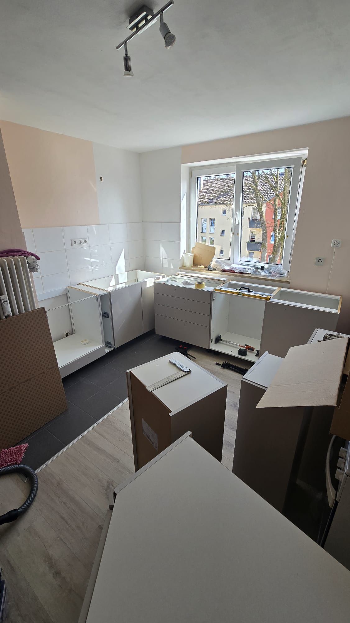Partially assembled kitchen cabinets inside a sunlit room with light wood and dark tiled flooring, a window overlooks neighboring buildings.