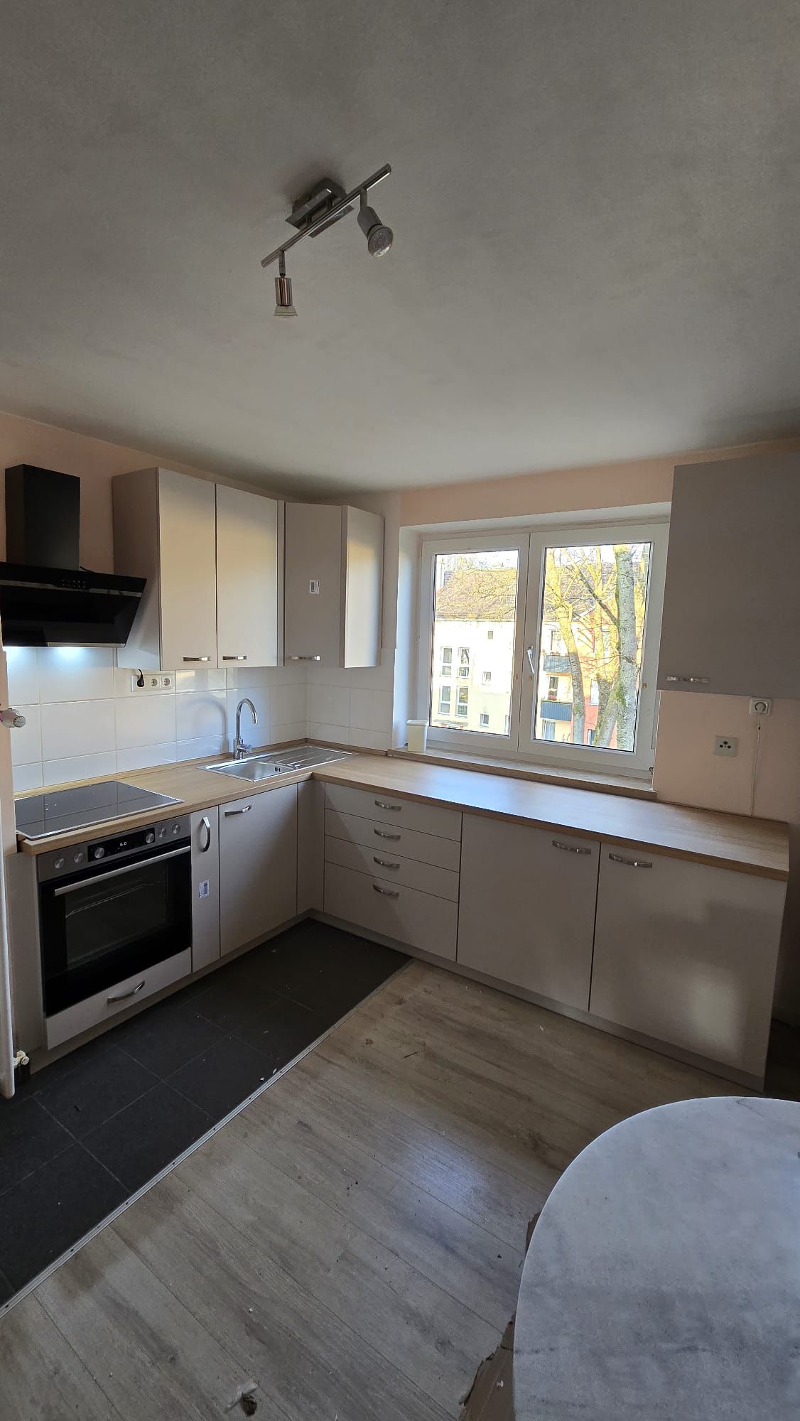 Modern kitchen with light wood countertops, beige cabinets, built-in oven, electric cooktop, stainless steel sink under a large window, and gray flooring.