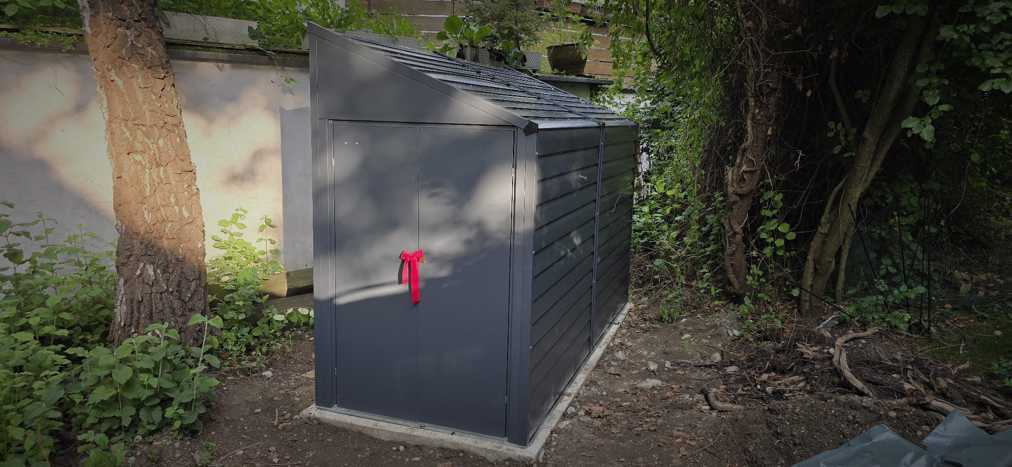 Dark gray metal garden shed with a slanted roof and a red ribbon tied on the front doors, surrounded by trees and plants.