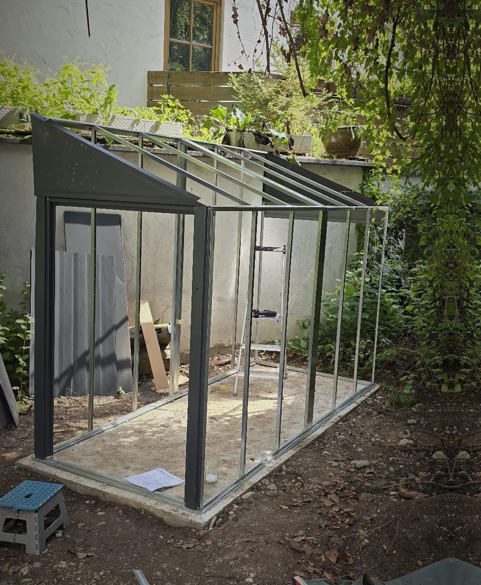 Partially assembled metal-framed greenhouse structure in a garden area with plants and a window in the background.