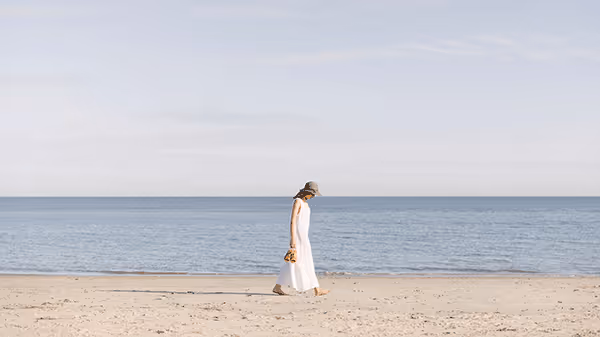 A woman in a long white dress walks barefoot along a quiet sandy beach, holding her sandals. The sky is pale and calm above the still, blue ocean stretching across the horizon.