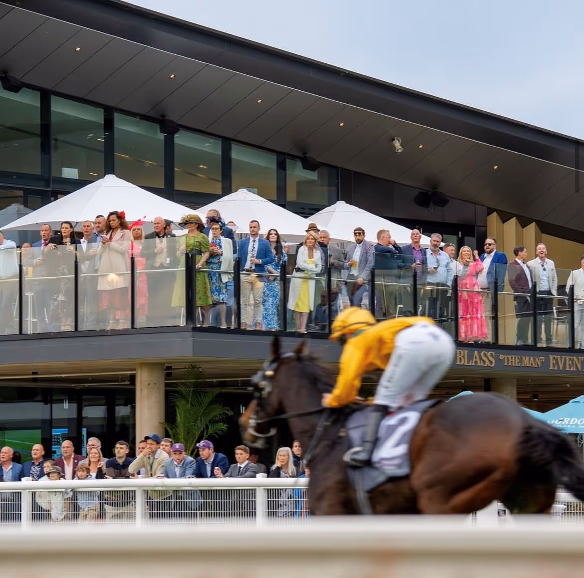 A horse race in motion with a jockey in yellow riding past a stylish crowd gathered on a modern grandstand balcony, dressed in race-day attire under white umbrellas.