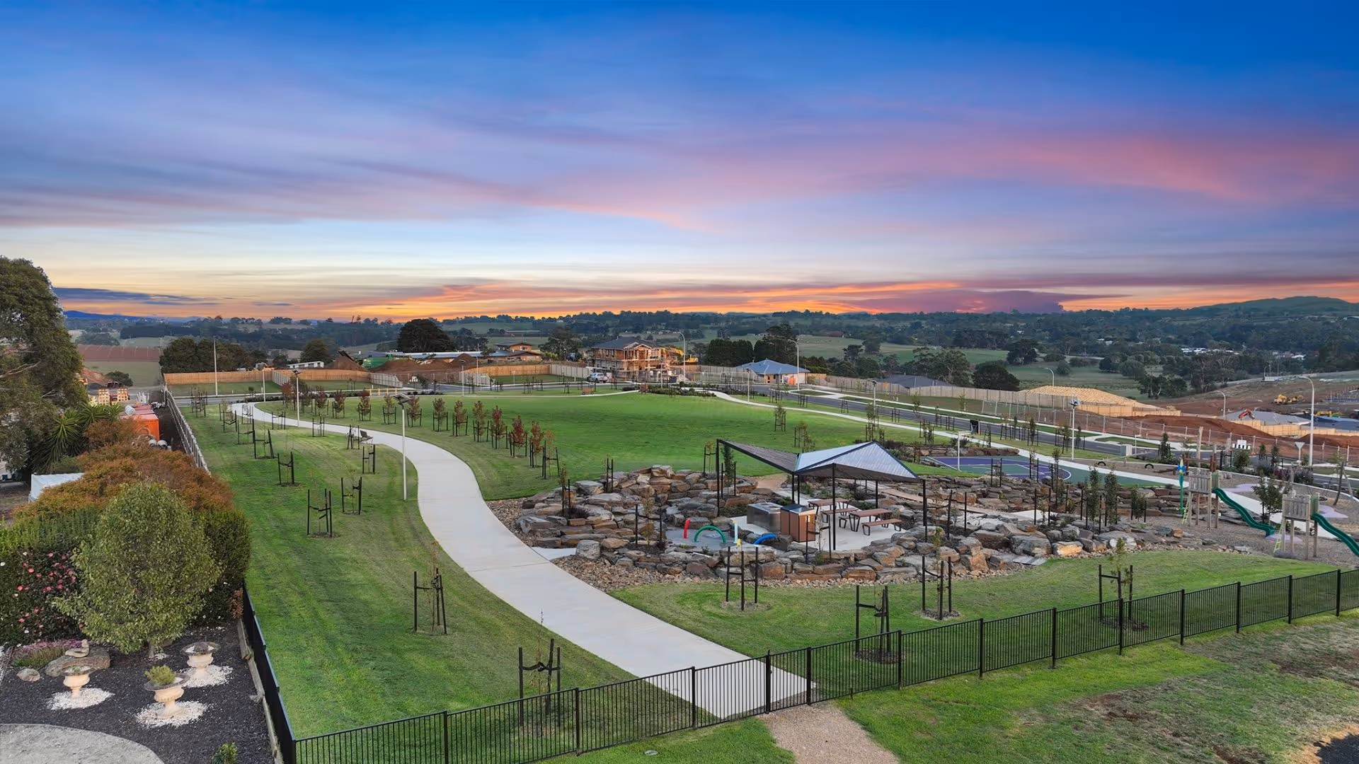 A large community park at sunset with winding paths, young trees, rock play areas and views of nearby houses under a colourful sky.