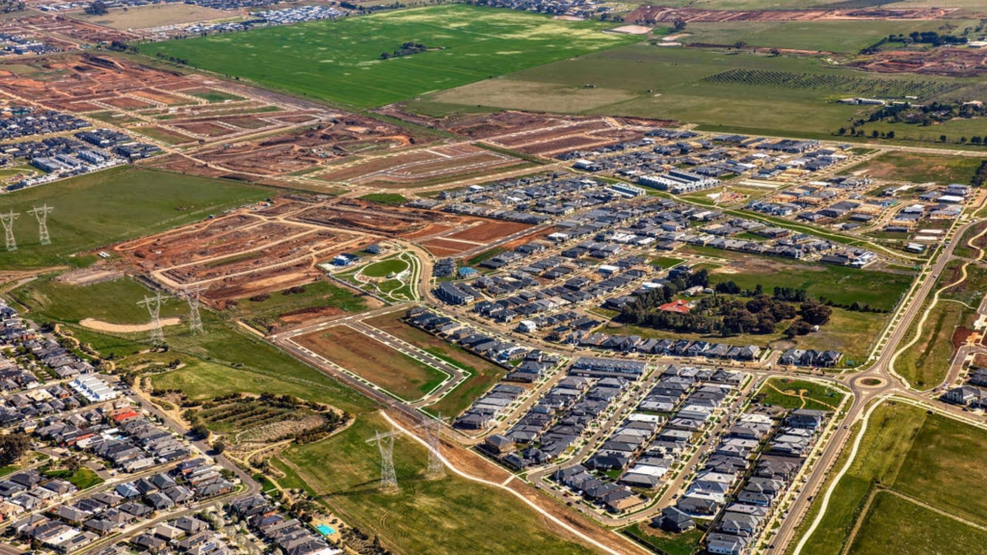 Aerial view of a large residential development under construction, with new roads, housing blocks, and green fields surrounding established neighbourhoods.