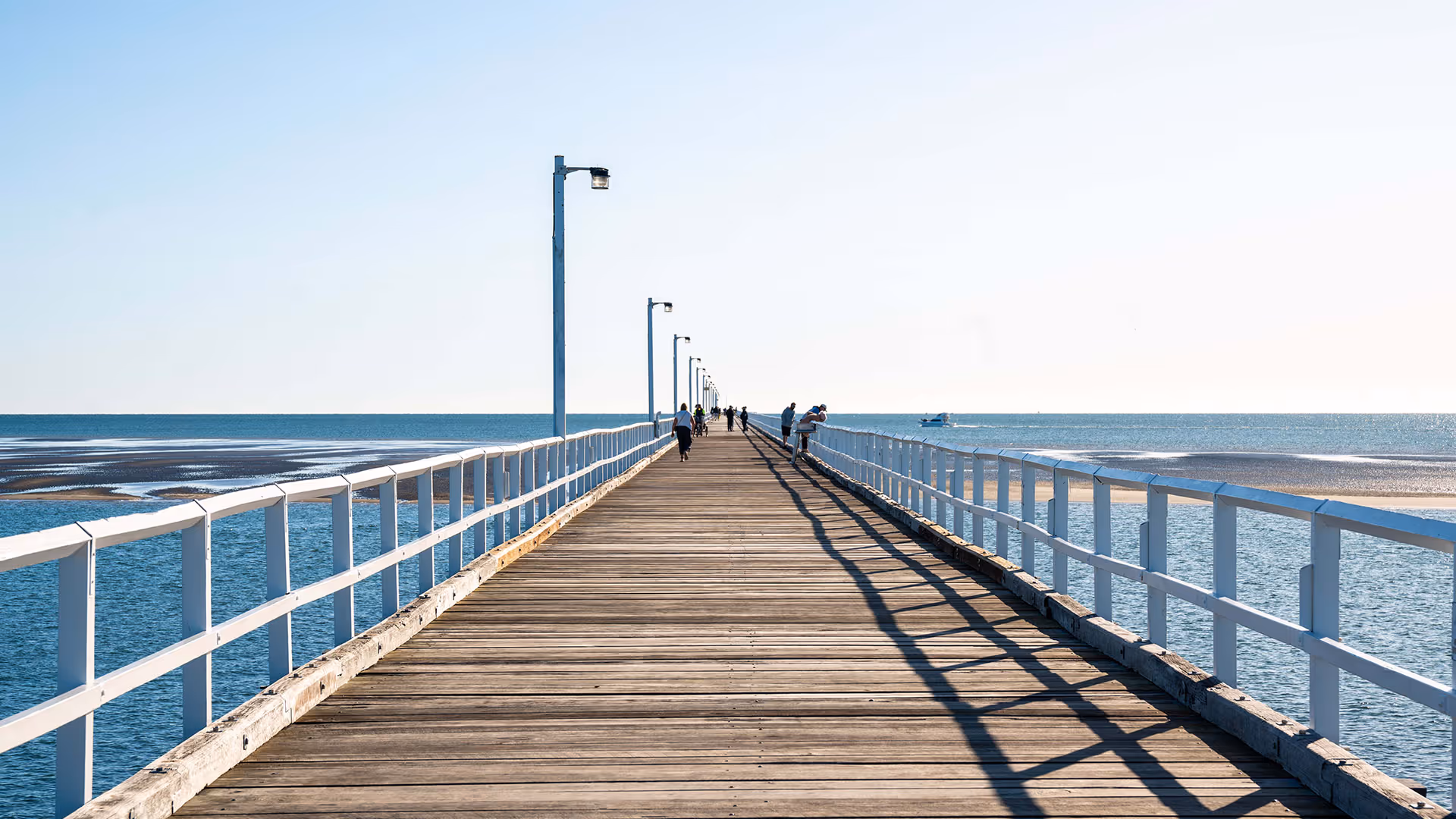 Long wooden pier stretching over calm blue water with people walking toward the horizon.