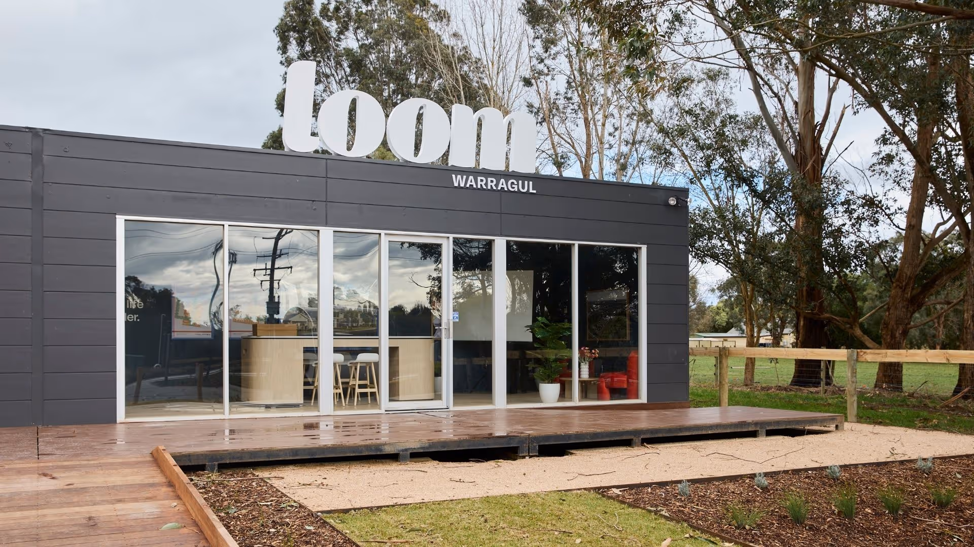 A modern dark-grey display suite with large glass windows and a bright white “loom Warragul” sign on top, surrounded by tall gum trees and a wooden deck at the entrance.