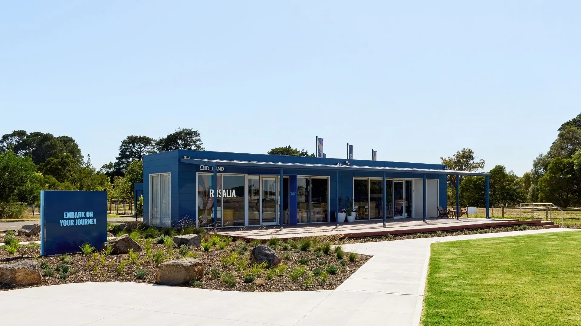 A modern blue sales office building surrounded by landscaped gardens, featuring large glass windows, potted plants, and a sign encouraging visitors to embark on their journey.