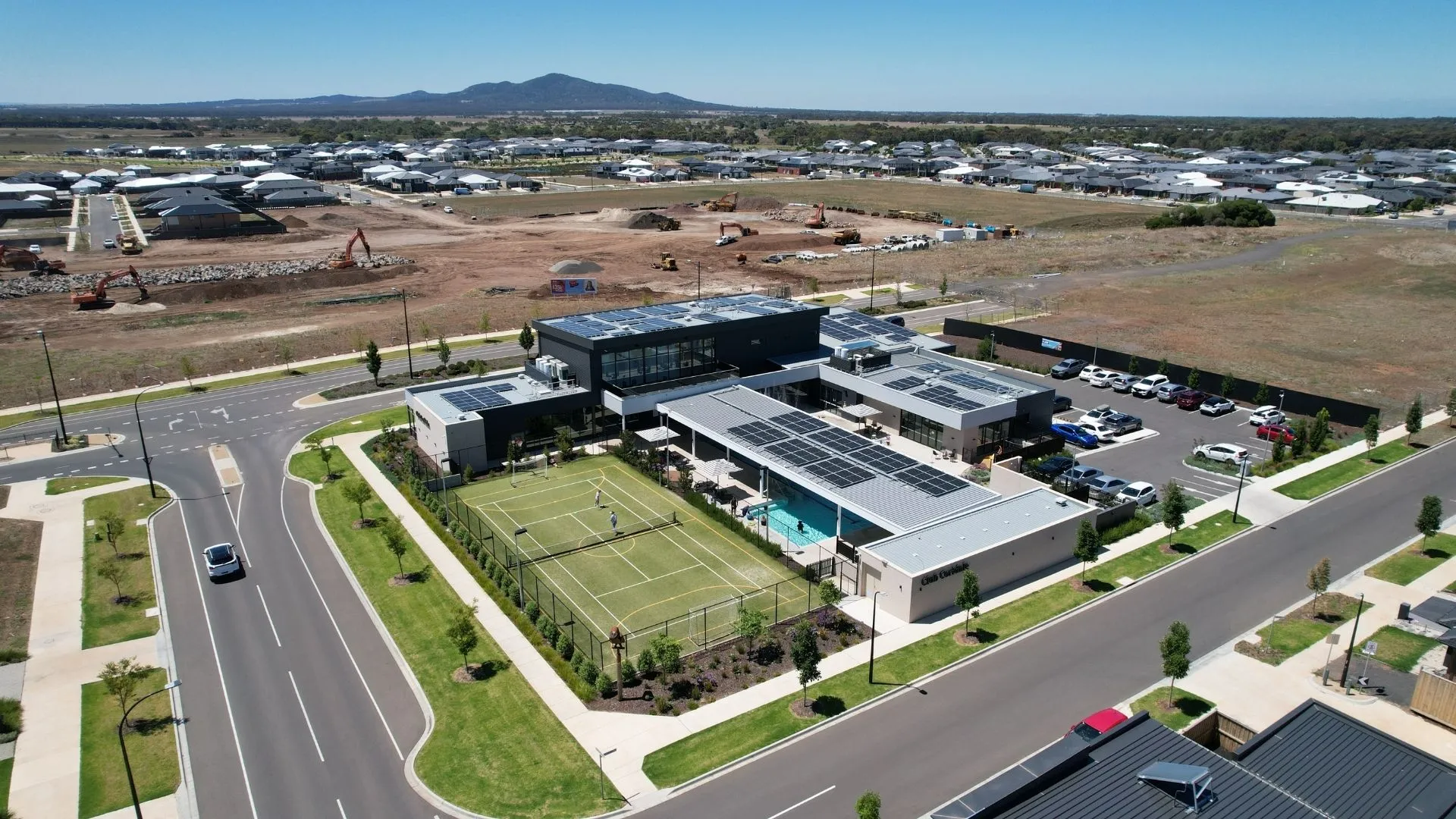 Aerial view of a modern community centre with tennis courts and pool beside new housing development.