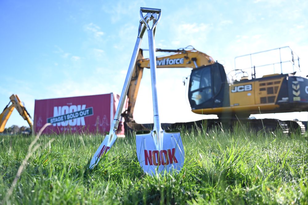Two Nook-branded ceremonial shovels stand upright in the grass at The Nook Colac, with a JCB excavator and 'Stage 1 Sold Out' hoarding in the background.