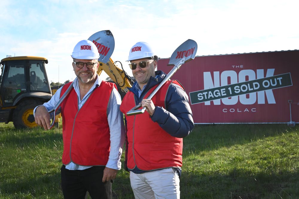 Two men in red Nook-branded hi-vis vests and white hard hats hold ceremonial shovels over their shoulders at The Nook Colac construction site, with an excavator and 'Stage 1 Sold Out' signage behind them.