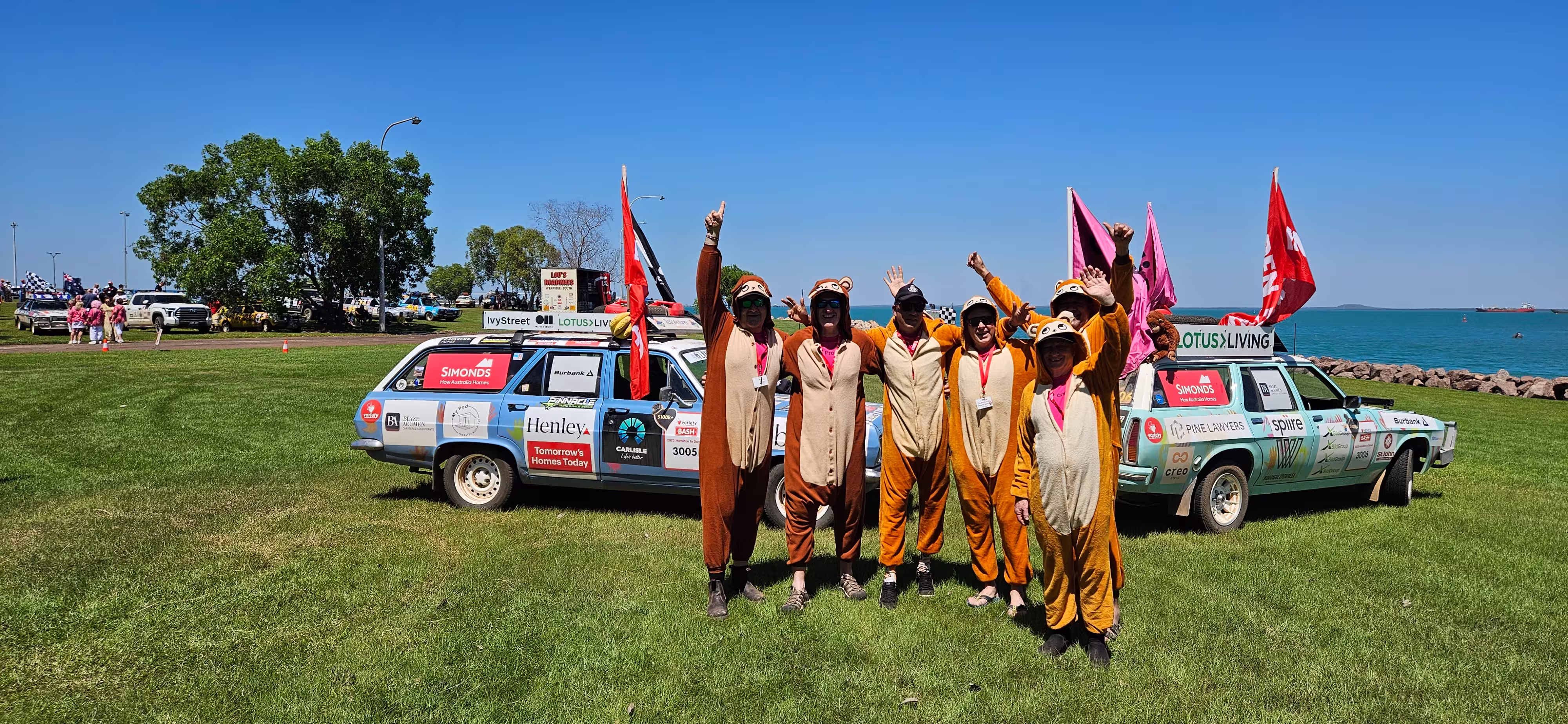 Team members in monkey costumes standing beside two sponsor-branded rally cars on a grassy waterfront during the Variety Bash charity event.