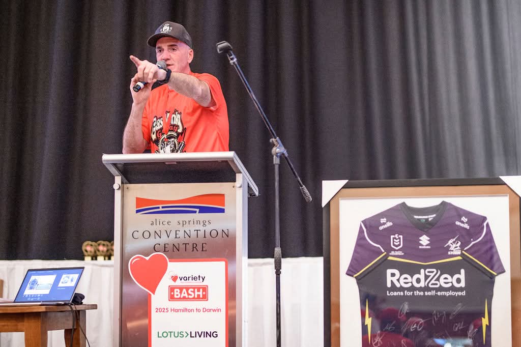 Speaker at a podium displaying Variety Bash branding at the Alice Springs Convention Centre, with a framed signed sports jersey beside the stage.