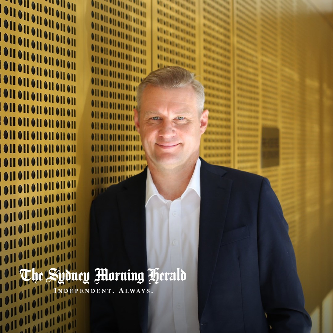 Oliver Hume Chief Economist Matt Bell in a navy blazer and white shirt, leaning against a gold perforated wall, with The Sydney Morning Herald logo